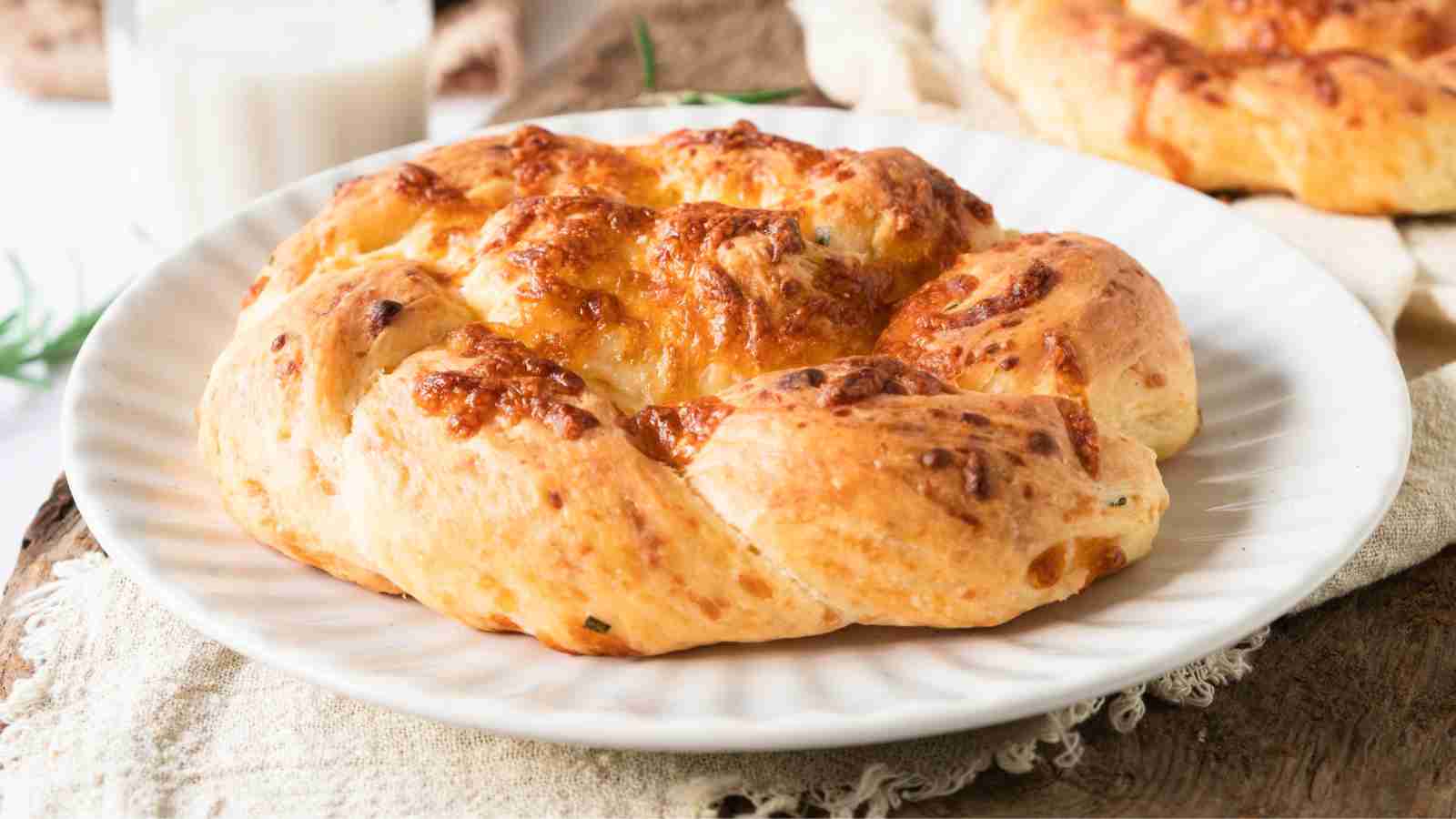 A round, golden-brown cheese bread with a braided pattern sits on a white plate, with another bread and a glass of milk in the background.