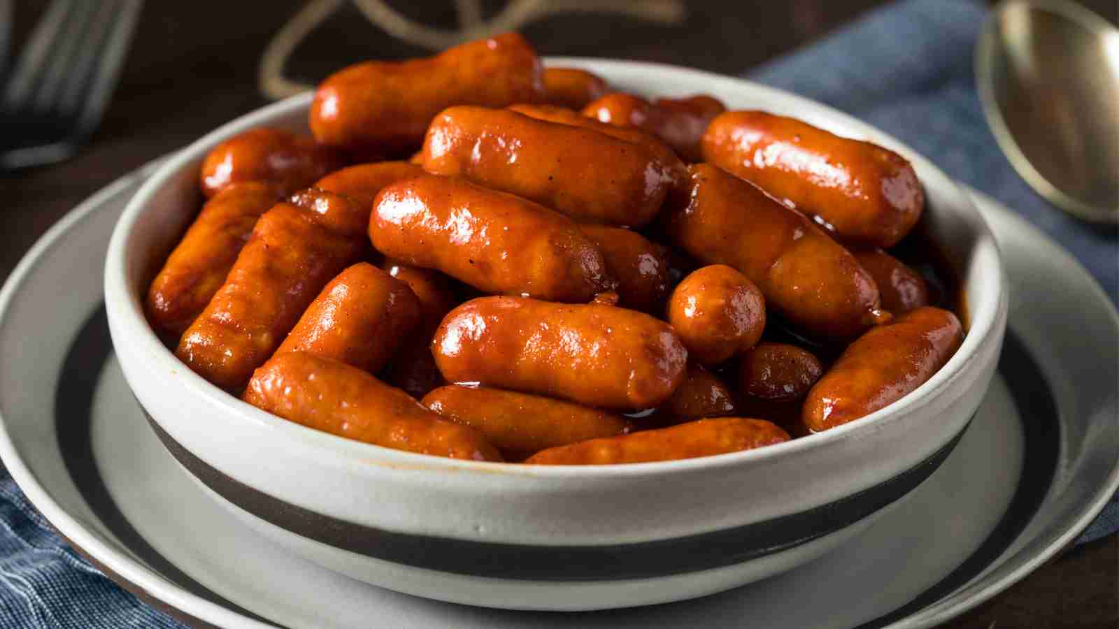 A white bowl filled with cocktail sausages coated in a glossy sauce, sitting on a plate with a spoon nearby.