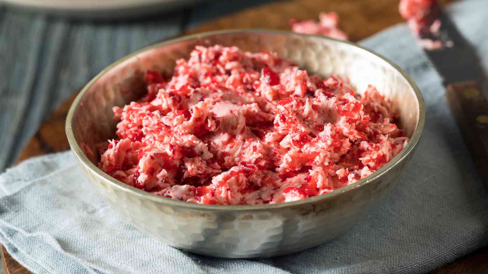 A metal bowl filled with a mixture of crushed cranberries and creamy ingredients sits on a blue cloth.
