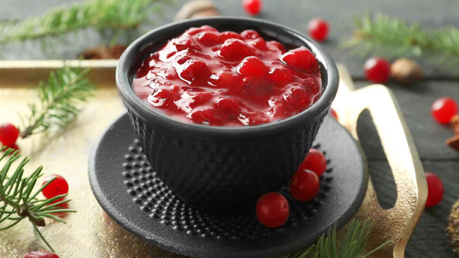 A black bowl filled with cranberry sauce sits on a matching plate, surrounded by a few loose cranberries and pine branches on a tray.
