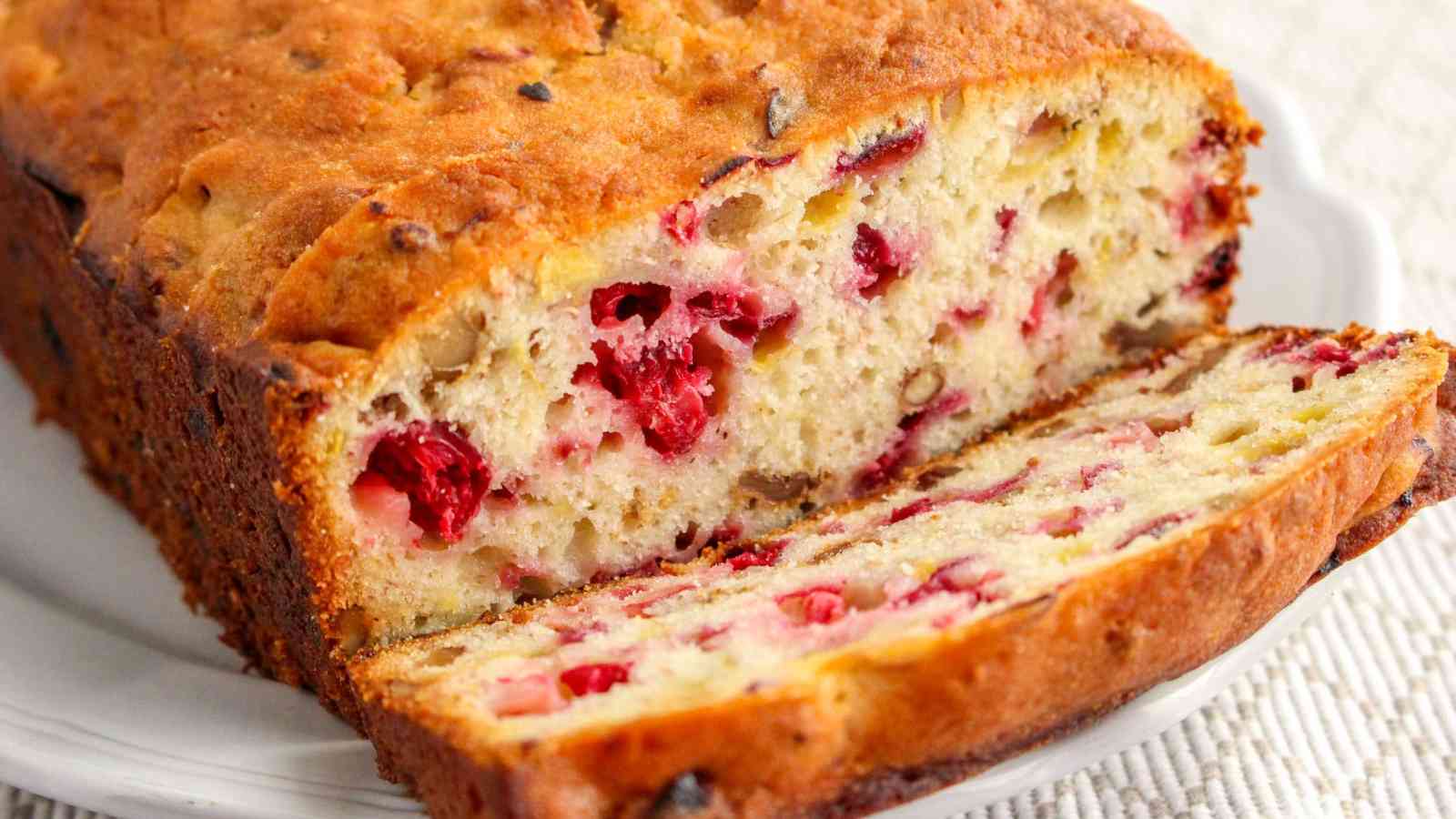 A loaf of cranberry bread with one slice cut, showing a moist, dense interior with visible cranberries throughout.