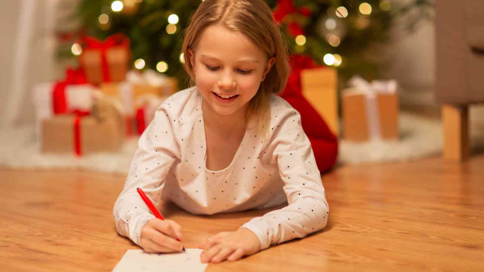 A young girl lies on the floor writing with a red pen on paper, with a Christmas tree and wrapped presents in the background.