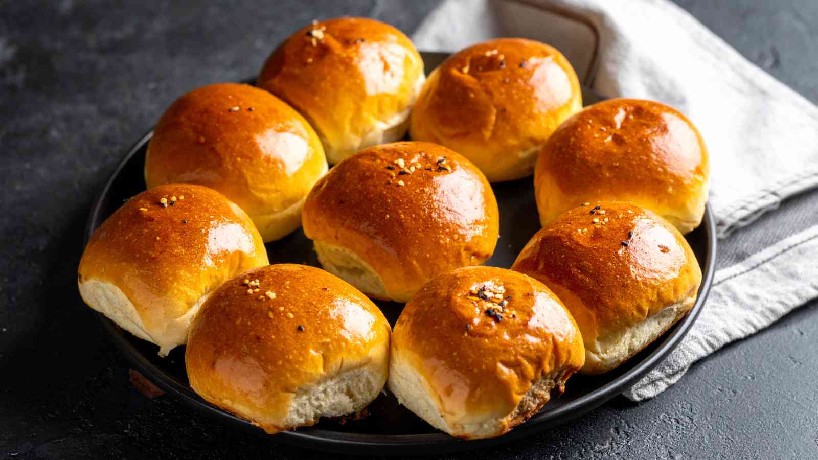 A round black plate holds nine shiny, golden-brown bread rolls, some topped with sesame seeds, on a dark surface with a folded beige napkin nearby.
