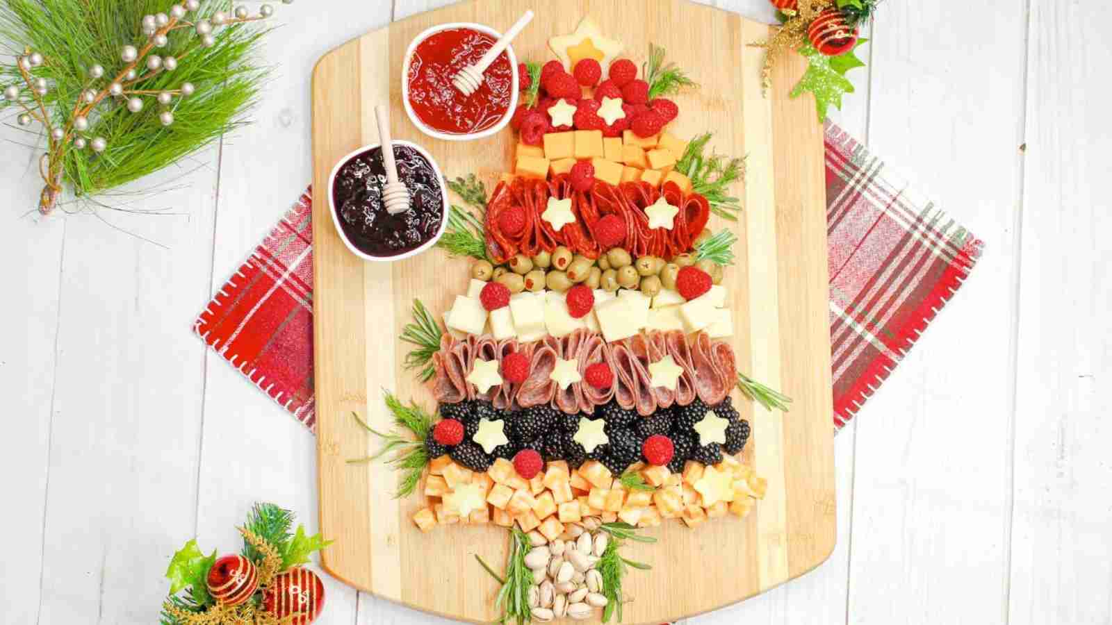 A Christmas Tree Charcuterie board, artfully arranged on a wooden board, features cheese, meats, olives, and berries with decorative star-shaped pieces. Two small bowls of jam sit at the top, surrounded by festive decorations for a delightful holiday display.