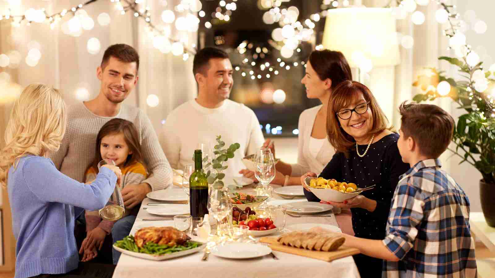 A group of six people, including two children, sit around a dinner table with food and drinks, smiling and talking in a warmly lit, decorated room.
