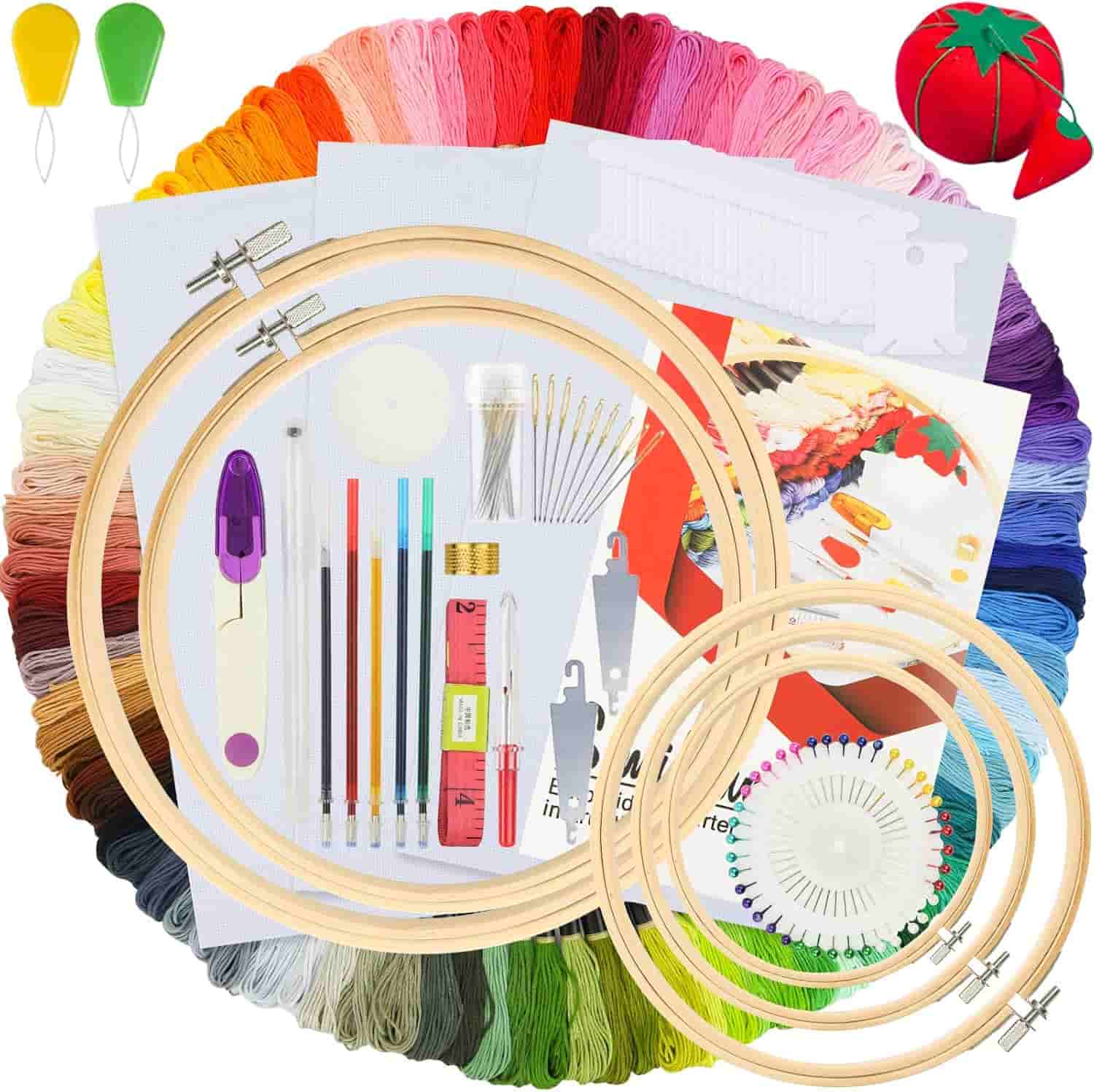 Embroidery kit with colored threads, wooden hoops, needles, needle threaders, threader tool, scissors, and a red pincushion arranged in a circular layout.