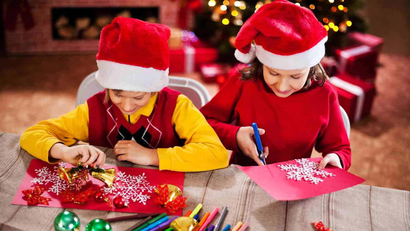Two children wearing Santa hats sit at a table making Christmas crafts with paper, decorations, and markers; a decorated Christmas tree is in the background.