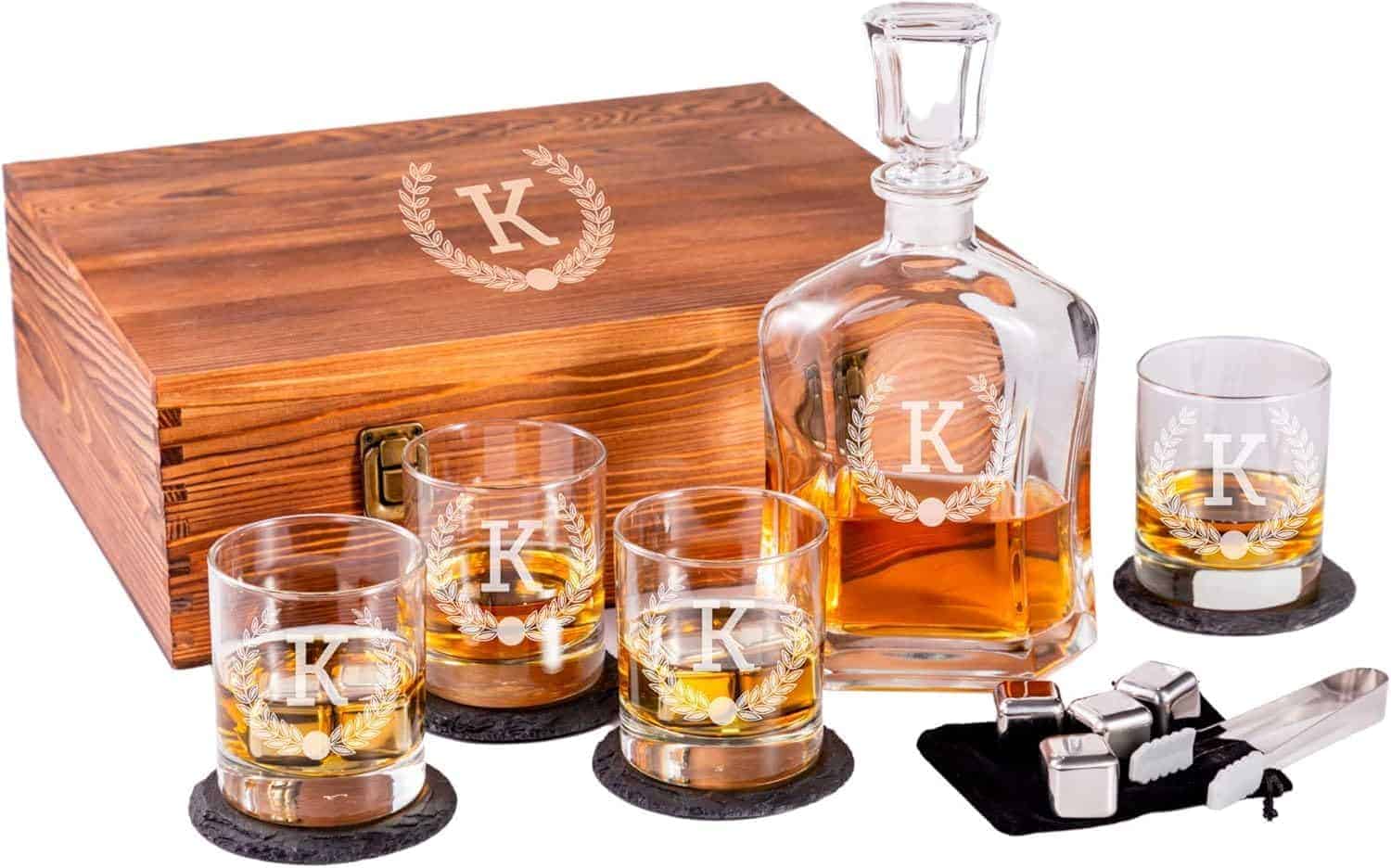 A whiskey decanter set with four glasses, each engraved with the letter K, coasters, a wooden box, whiskey stones, and metal tongs.