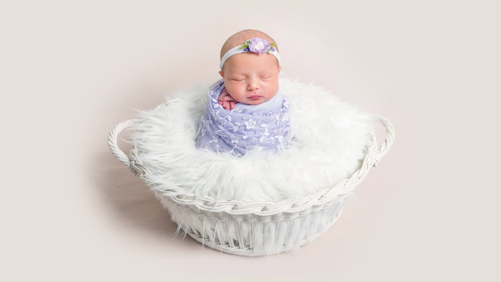 Newborn baby wrapped in a lavender blanket and floral headband, sleeping in a white, fluffy-lined basket against a light background.