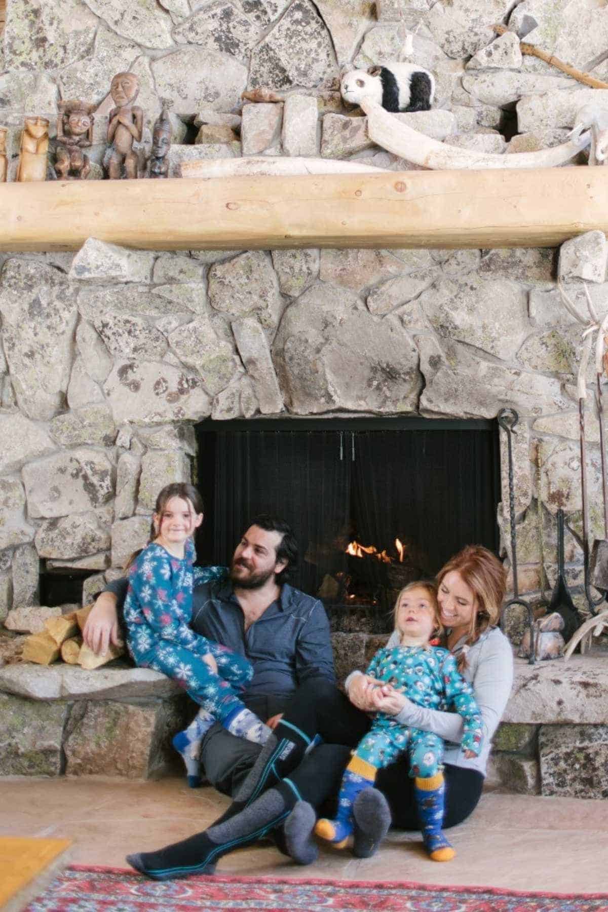 A family of four in pajamas sits together on a stone hearth in front of a lit fireplace, with animal figurines and wood on the mantel above.