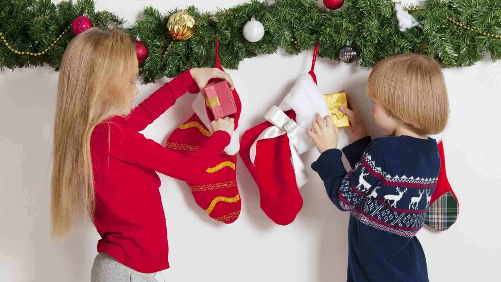 Two children place gifts into Christmas stockings hanging from a decorated garland.