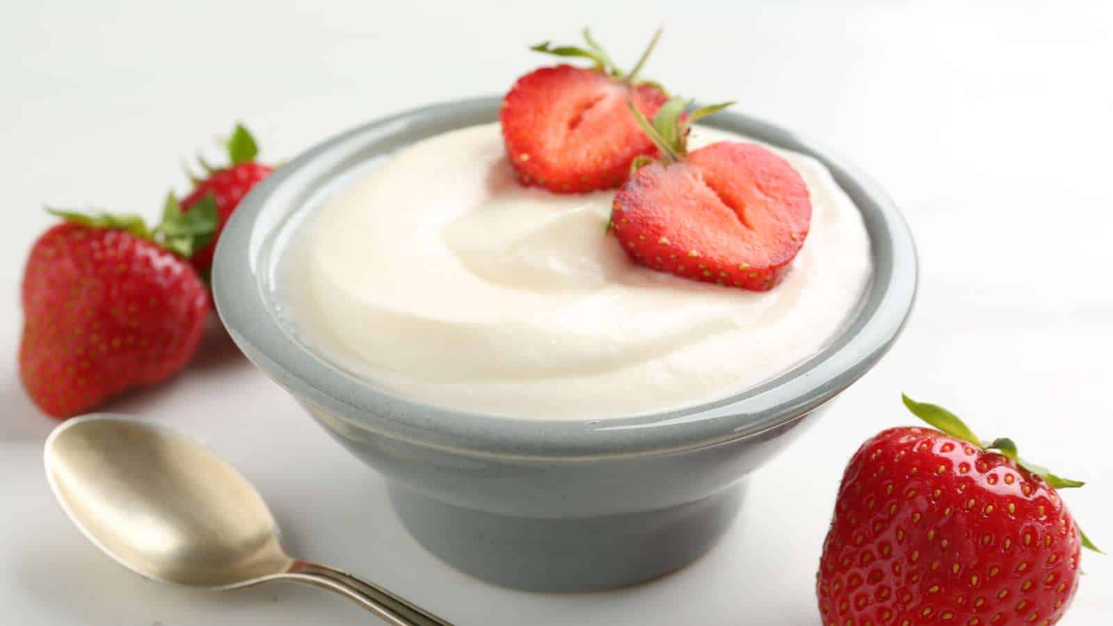 A bowl of plain yogurt topped with sliced strawberries, with whole strawberries and a spoon beside it on a white surface.