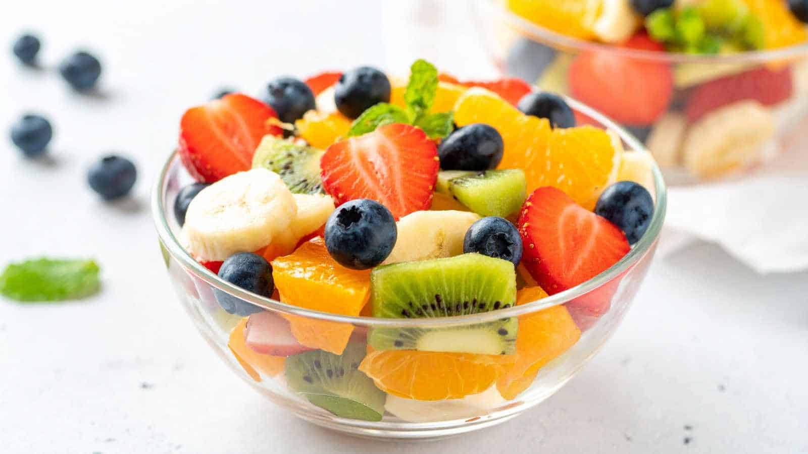 A clear glass bowl filled with a colorful fruit salad containing strawberries, blueberries, kiwi, banana slices, orange segments, and mint leaves.