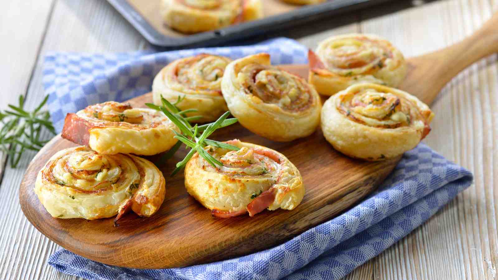 A wooden board with several puff pastry pinwheels filled with ham and cheese, garnished with rosemary, on a blue checkered cloth.