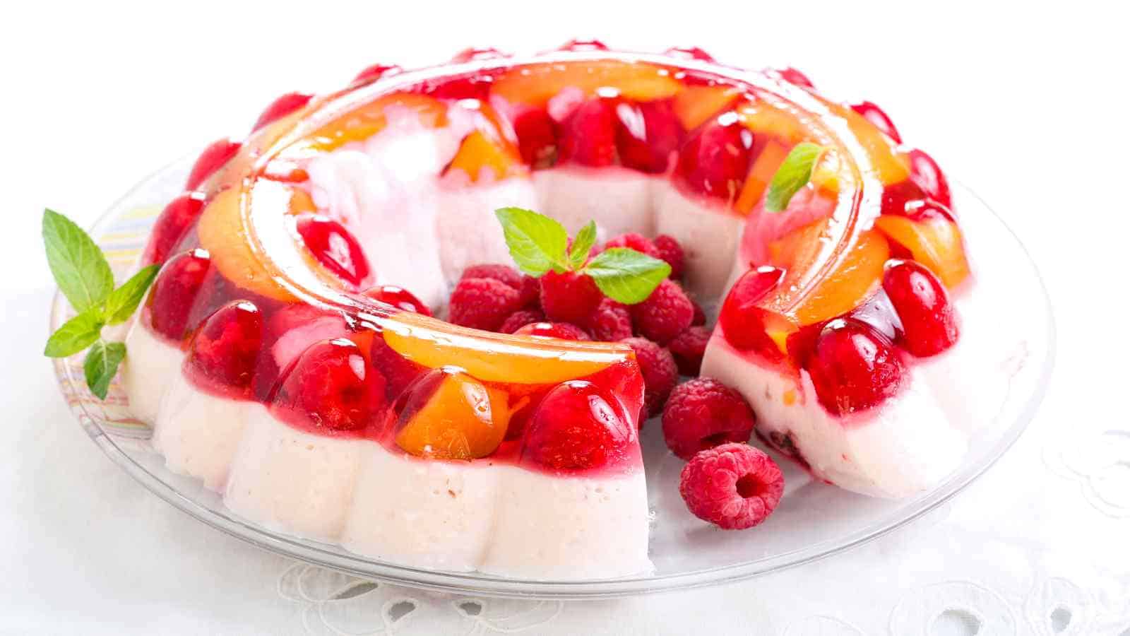 A gelatin and cream dessert ring with embedded strawberries, peaches, and raspberries, garnished with mint leaves and fresh raspberries on a glass plate.