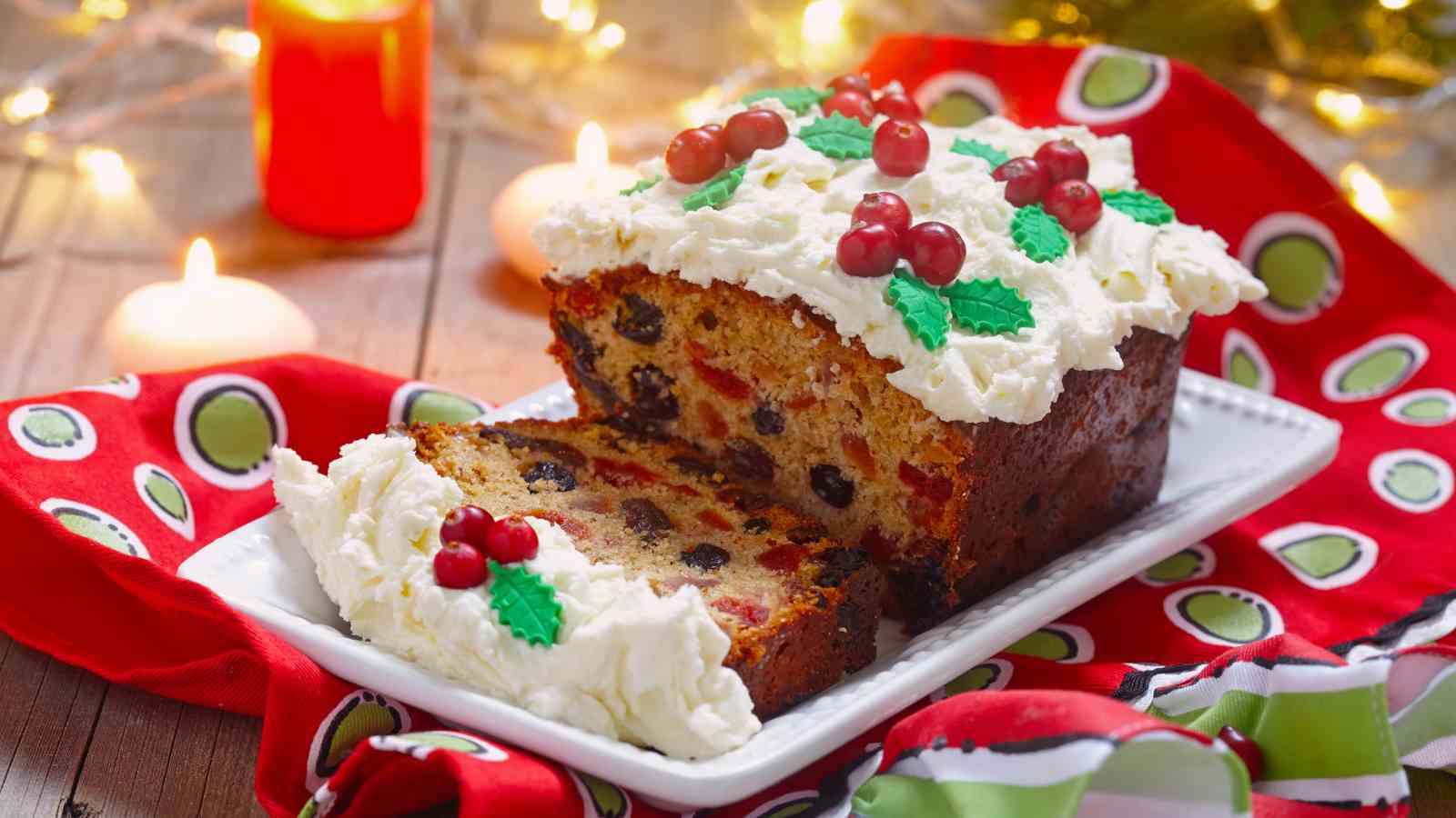 A sliced fruitcake topped with white frosting, red berries, and green leaf decorations sits on a white platter, surrounded by candles and holiday lights.