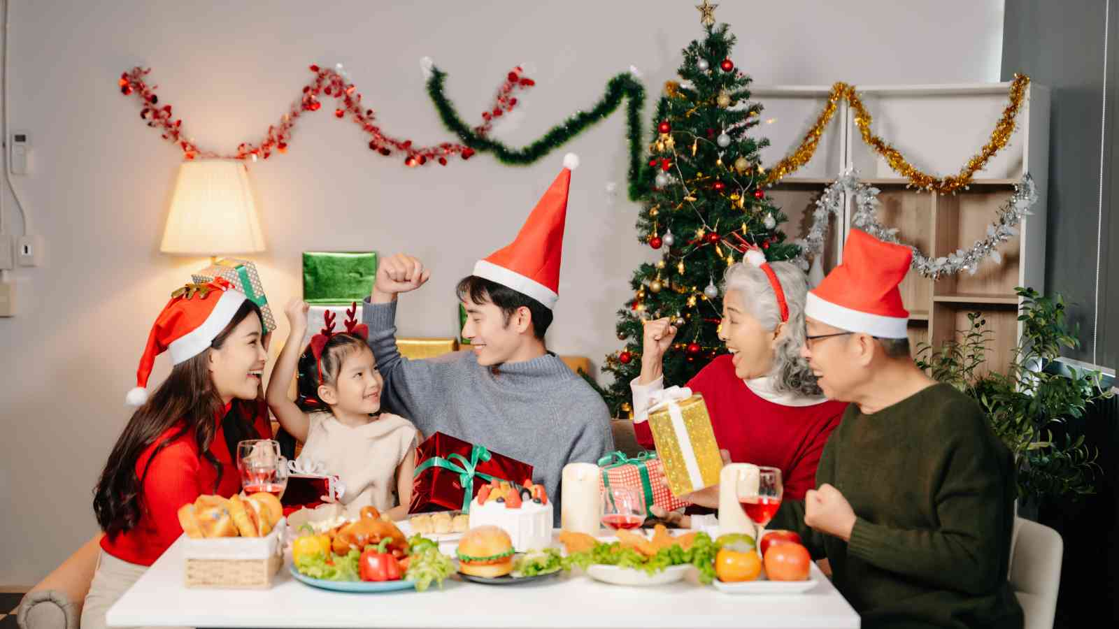 Five people wearing Santa hats sit at a table with food and gifts, smiling and raising their arms in celebration with Christmas decorations in the background.
