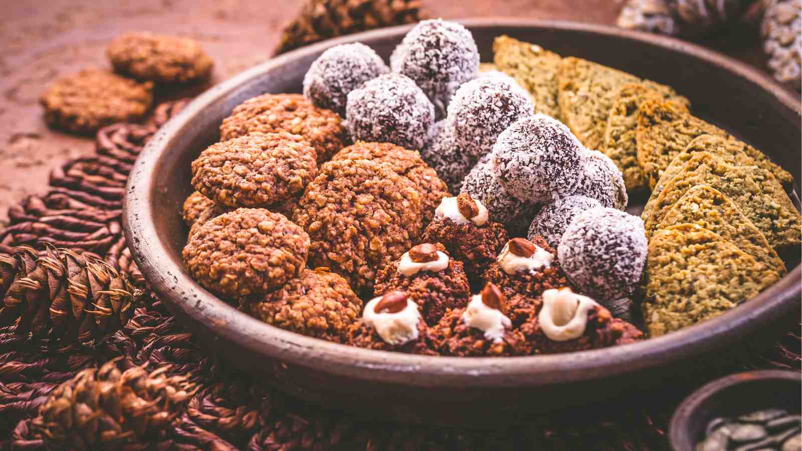 A ceramic plate filled with assorted cookies and treats, including coconut-covered balls, oatmeal cookies, green triangle-shaped cookies, and chocolate-topped bites.