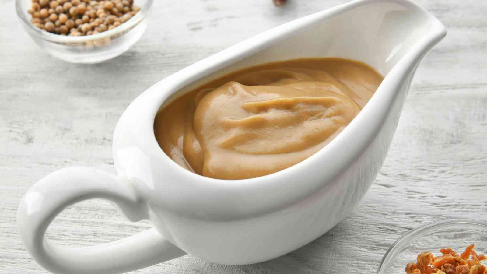 A white ceramic gravy boat filled with creamy brown gravy, placed on a light-colored surface with small bowls of spices nearby.