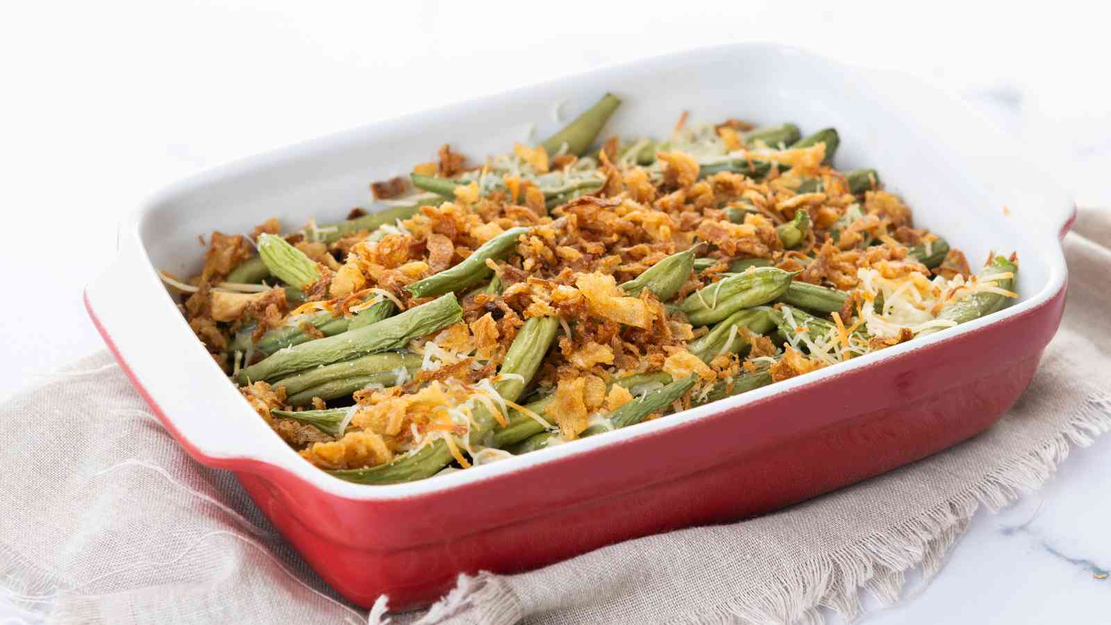 A red baking dish filled with green bean casserole, topped with crispy fried onions and shredded cheese, sits on a beige cloth.