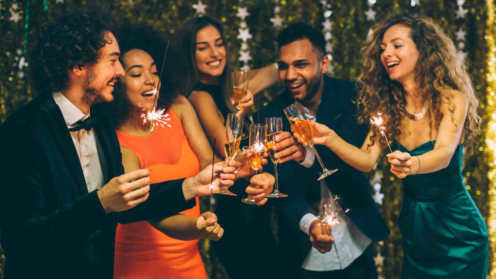 Five people dressed in formal attire hold sparklers and glasses of champagne, celebrating together in front of a gold tinsel backdrop.