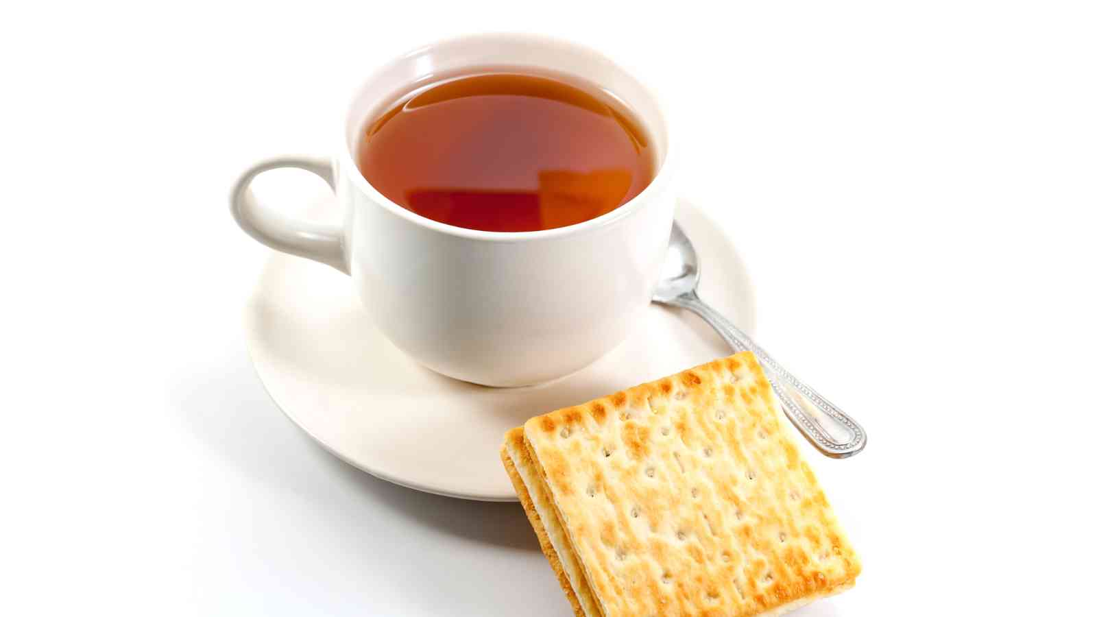 A white cup filled with tea on a saucer with a spoon, next to two square crackers, all set against a white background.