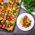 A sheet pan with roasted sausage, potatoes, broccoli, and bell peppers next to a white plate with a serving of the same food and a bunch of fresh parsley on the side.