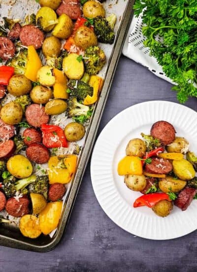 A sheet pan with roasted sausage, potatoes, broccoli, and bell peppers next to a white plate with a serving of the same food and a bunch of fresh parsley on the side.