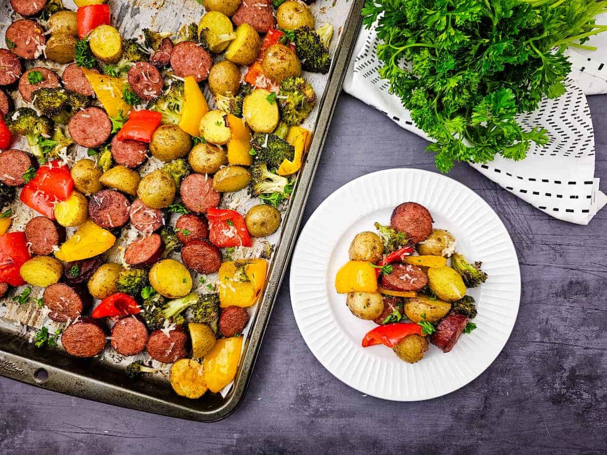 A sheet pan with roasted sausage, potatoes, broccoli, and bell peppers next to a white plate with a serving of the same food and a bunch of fresh parsley on the side.