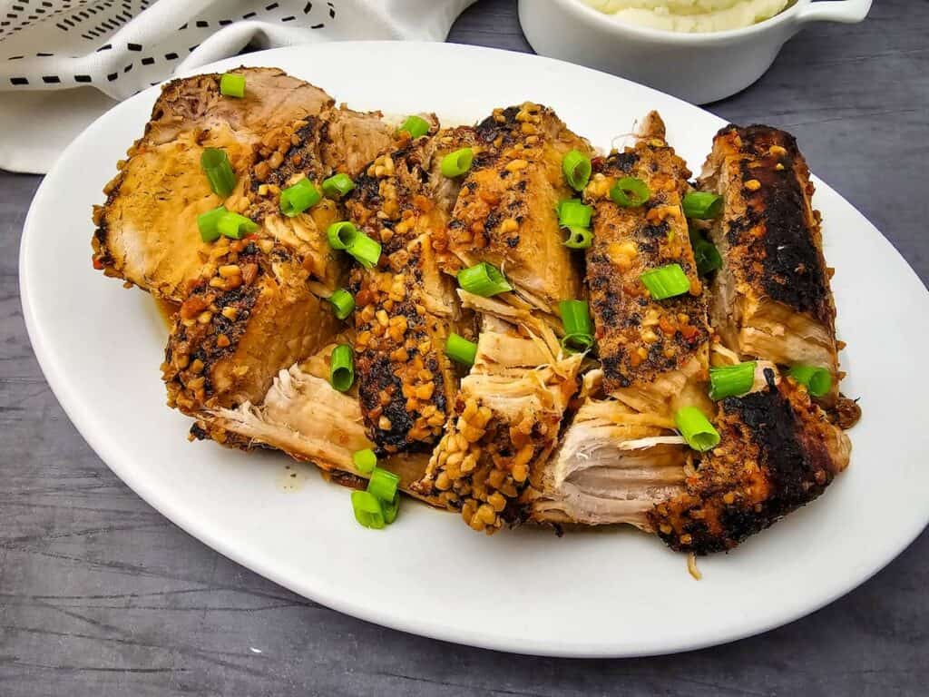 Sliced, seasoned pork roast garnished with chopped green onions on a white oval plate, set on a dark surface.