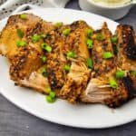 Sliced, seasoned pork roast garnished with chopped green onions on a white oval plate, set on a dark surface.