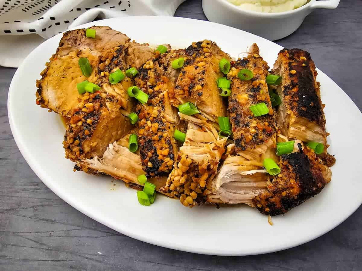 Sliced, seasoned pork roast garnished with chopped green onions on a white oval plate, set on a dark surface.