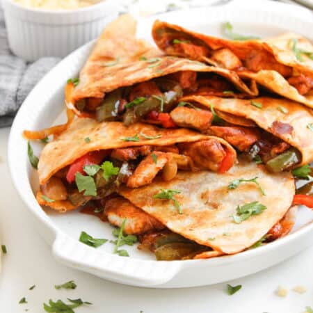 A plate of chicken quesadillas filled with sautéed peppers and onions, garnished with fresh cilantro, sits on a white surface.