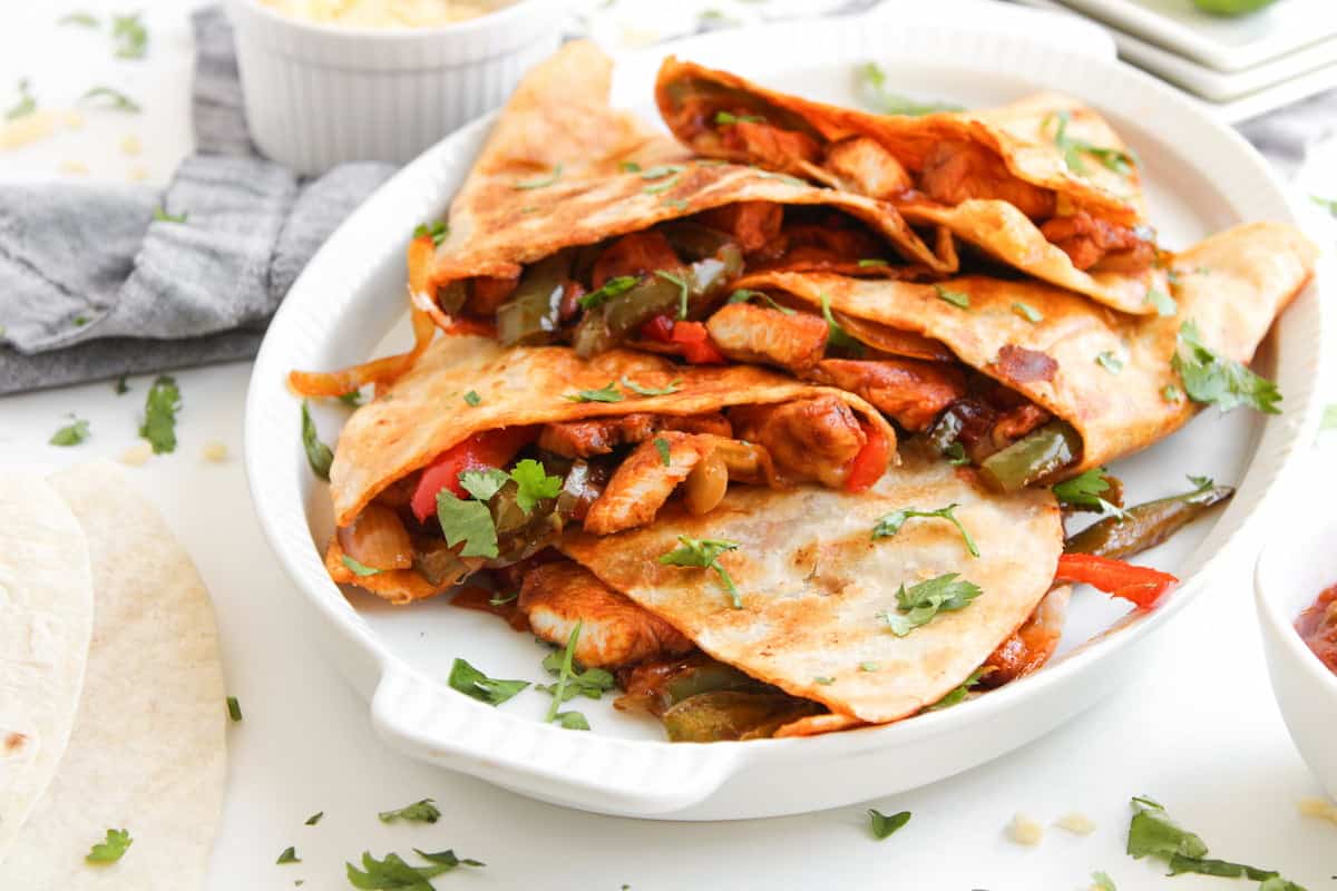 A plate of chicken quesadillas filled with sautéed peppers and onions, garnished with fresh cilantro, sits on a white surface.