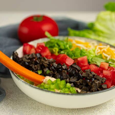 A bowl of layered cottage cheese dip topped with chopped tomatoes, shredded cheese, lettuce, olives, green onions, and a carrot stick.