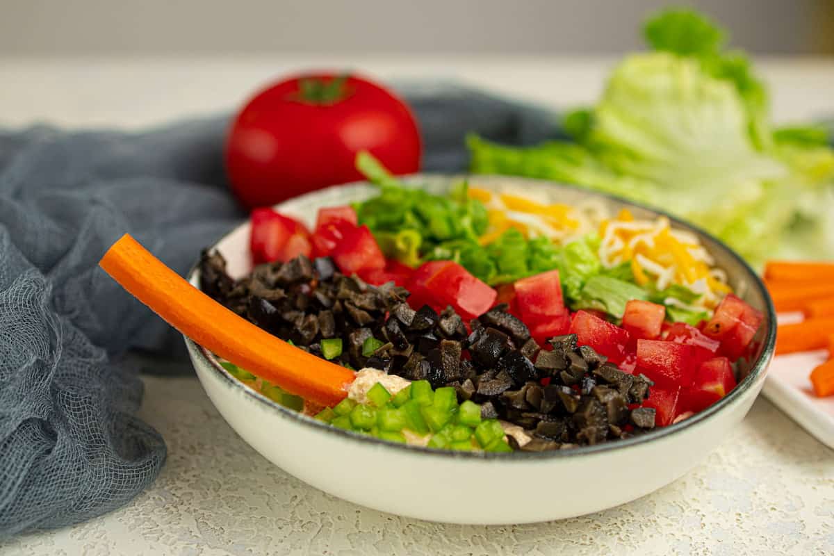 A bowl of salad with chopped tomatoes, black olives, green peppers, lettuce, shredded cheese, and a carrot stick, with a tomato and lettuce in the background.