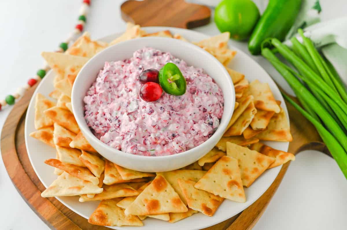 A bowl of creamy dip garnished with cranberries and a jalapeño slice is surrounded by chips on a white plate, with green onions and limes in the background.
