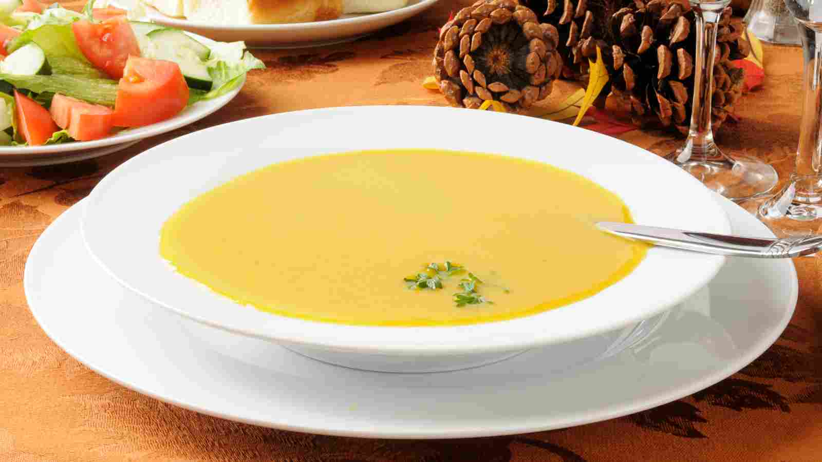 A bowl of creamy yellow soup garnished with herbs, placed on a white plate with a spoon, next to a salad and decorative pinecones on a brown tablecloth.