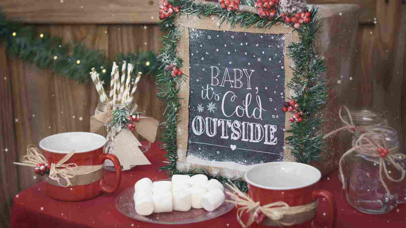 A festive table with two red mugs, marshmallows on a plate, and a chalkboard sign reading “Baby it’s cold outside,” decorated with greenery and berries.