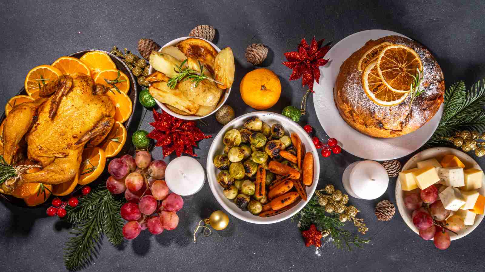 A table set with roasted chicken, sliced oranges, roasted potatoes, brussels sprouts, carrots, grapes, cheese cubes, a citrus-topped cake, candles, and festive holiday decorations.