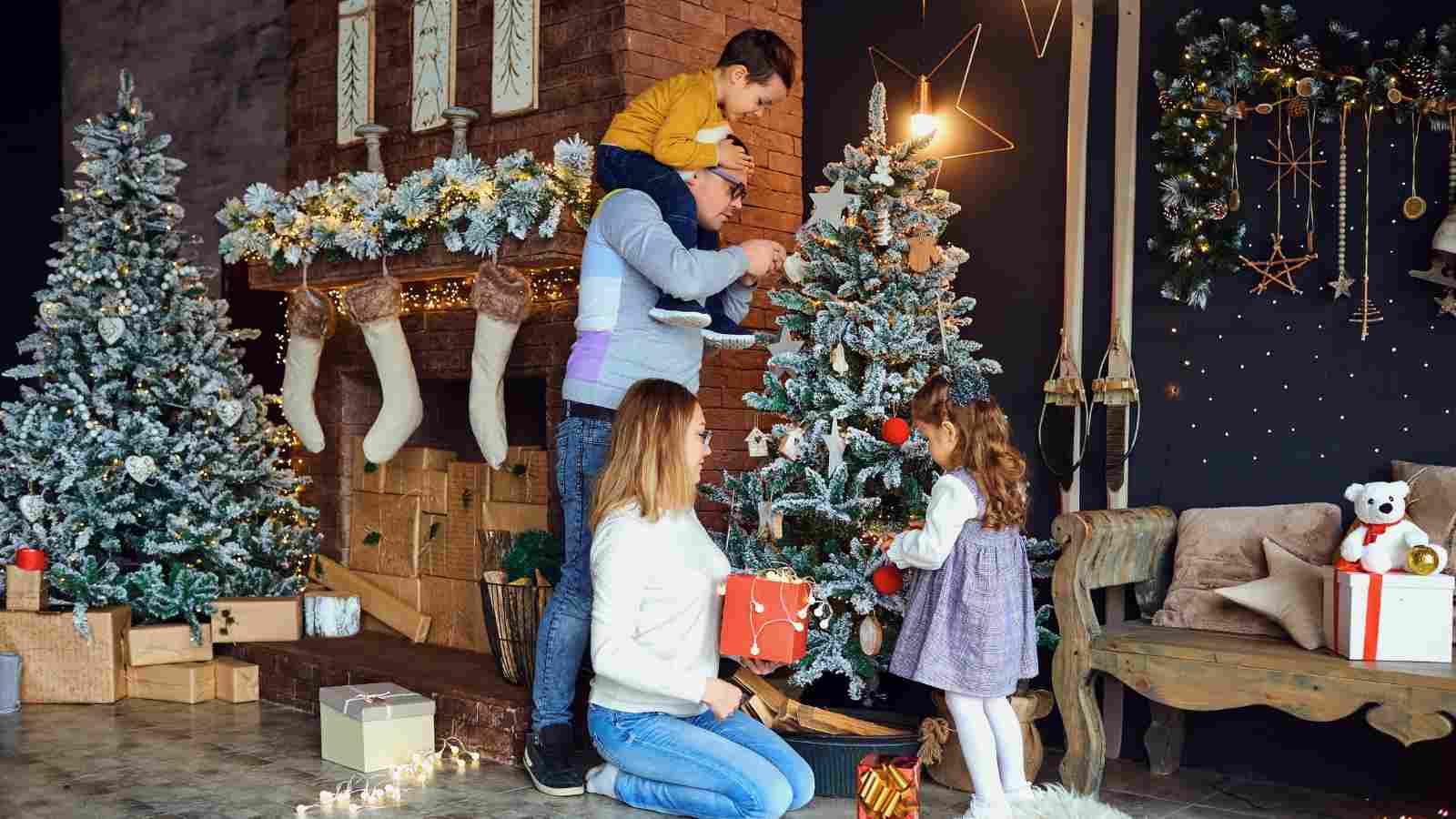 A family decorates a Christmas tree in a living room with a fireplace, stockings, wrapped gifts, and festive decorations.
