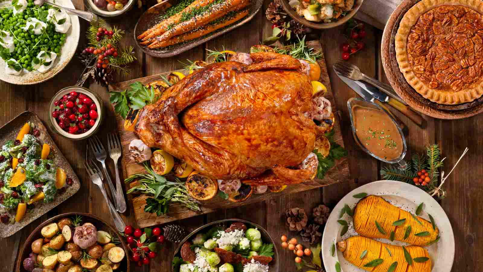 A roasted turkey is surrounded by various side dishes, including vegetables, salad, sauce, fresh herbs, and pies, all arranged on a wooden table.
