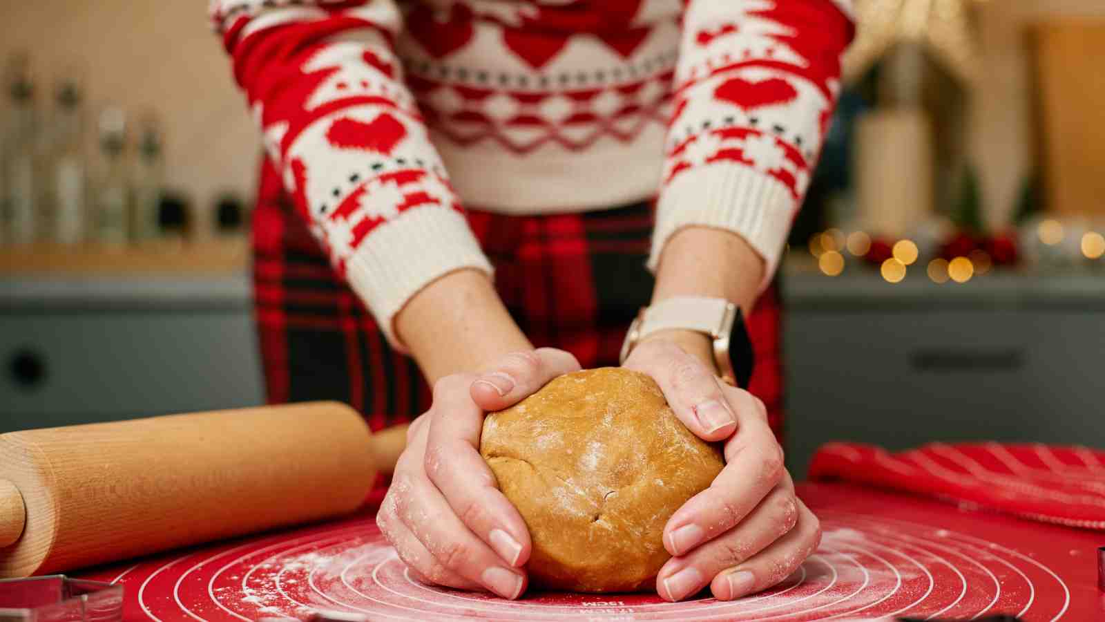 Person wearing a festive sweater kneads a ball of dough on a red pastry mat, with a rolling pin nearby.