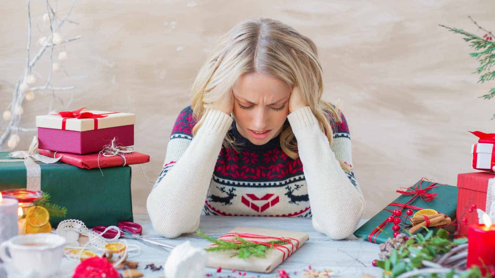 A woman in a holiday sweater sits at a table with wrapped gifts, resting her head in her hands and appearing stressed or overwhelmed.