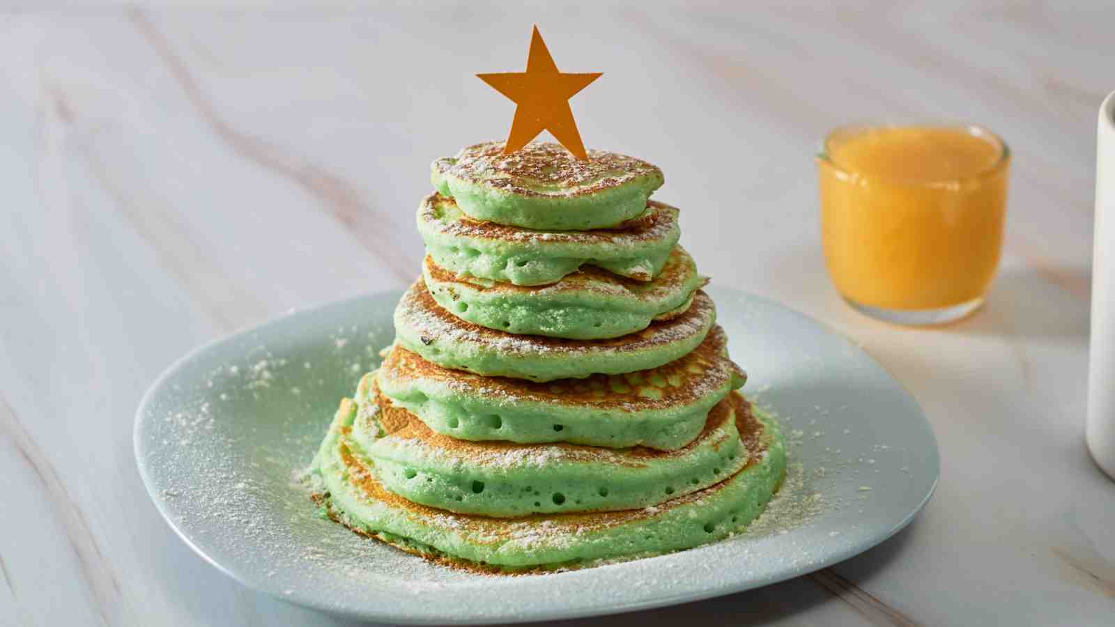 A stack of green pancakes arranged in a tree shape with a star topper, dusted with powdered sugar on a white plate. Orange juice and a white mug are in the background.