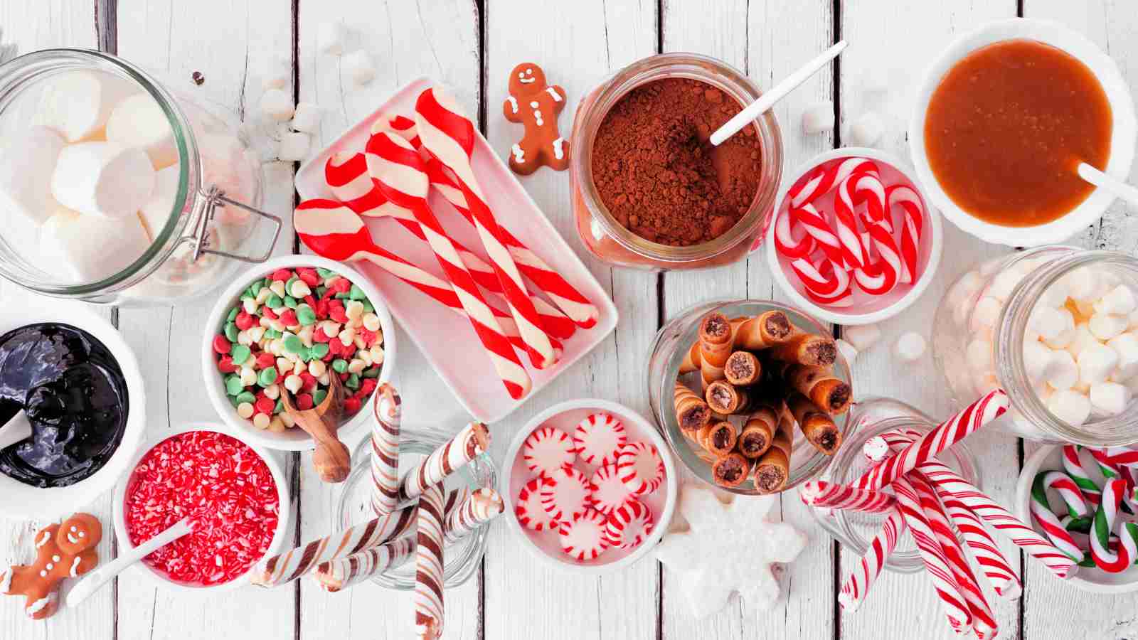 An assortment of holiday candies, cookies, jarred toppings, and marshmallows arranged on a white wooden surface.