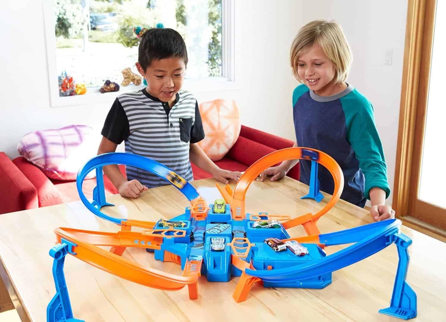 Two children play with a Hot Wheels track set on a wooden table in a brightly lit room with a red couch and a window in the background.
