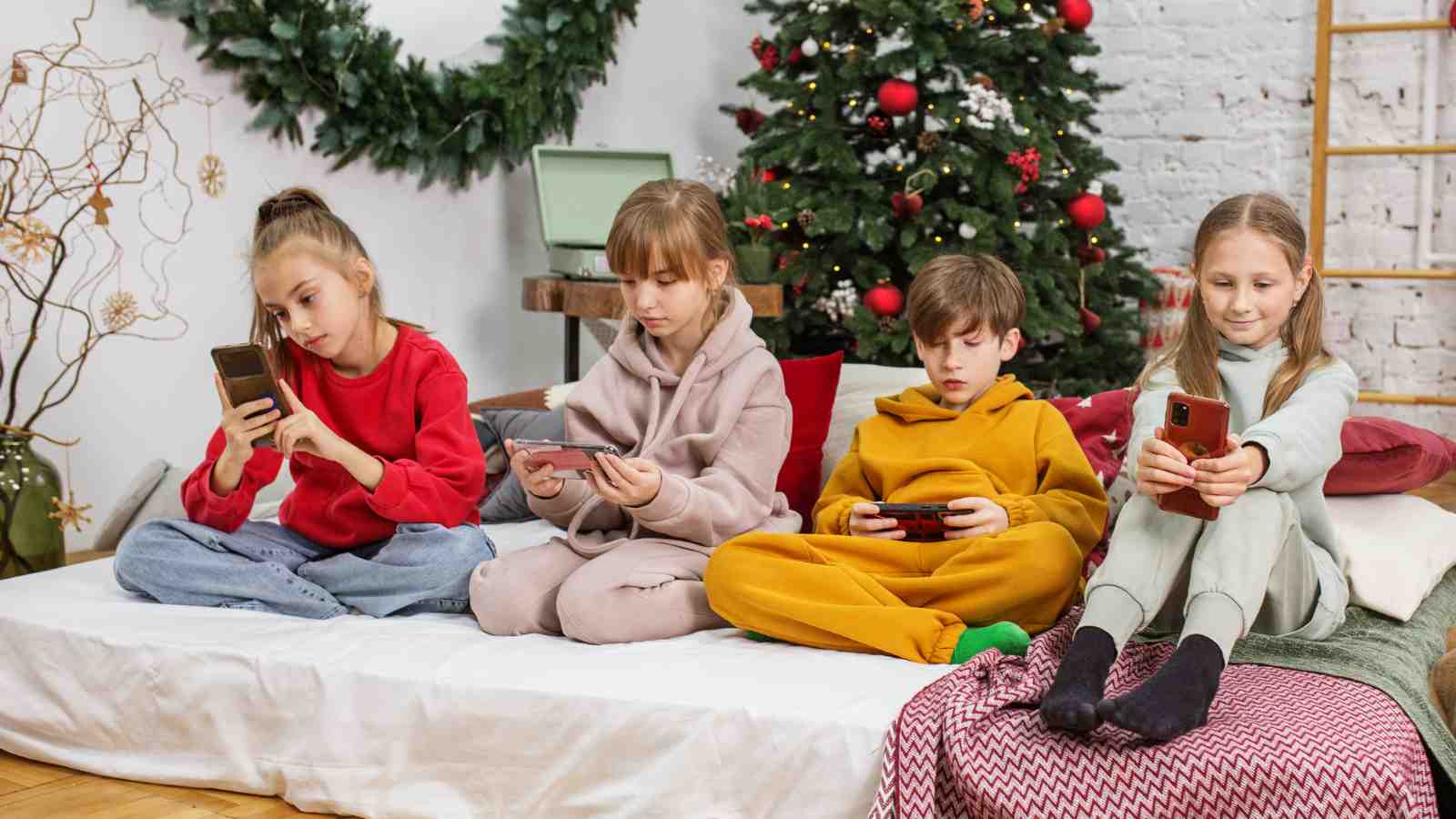 Four children in cozy outfits sit on a bed in a decorated room, each focused on their own handheld device, with a Christmas tree and wreath in the background.