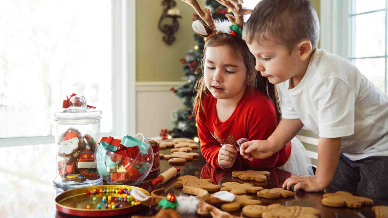 Two young children decorate gingerbread cookies at a table, surrounded by holiday decorations, candy, and a Christmas tree in the background.