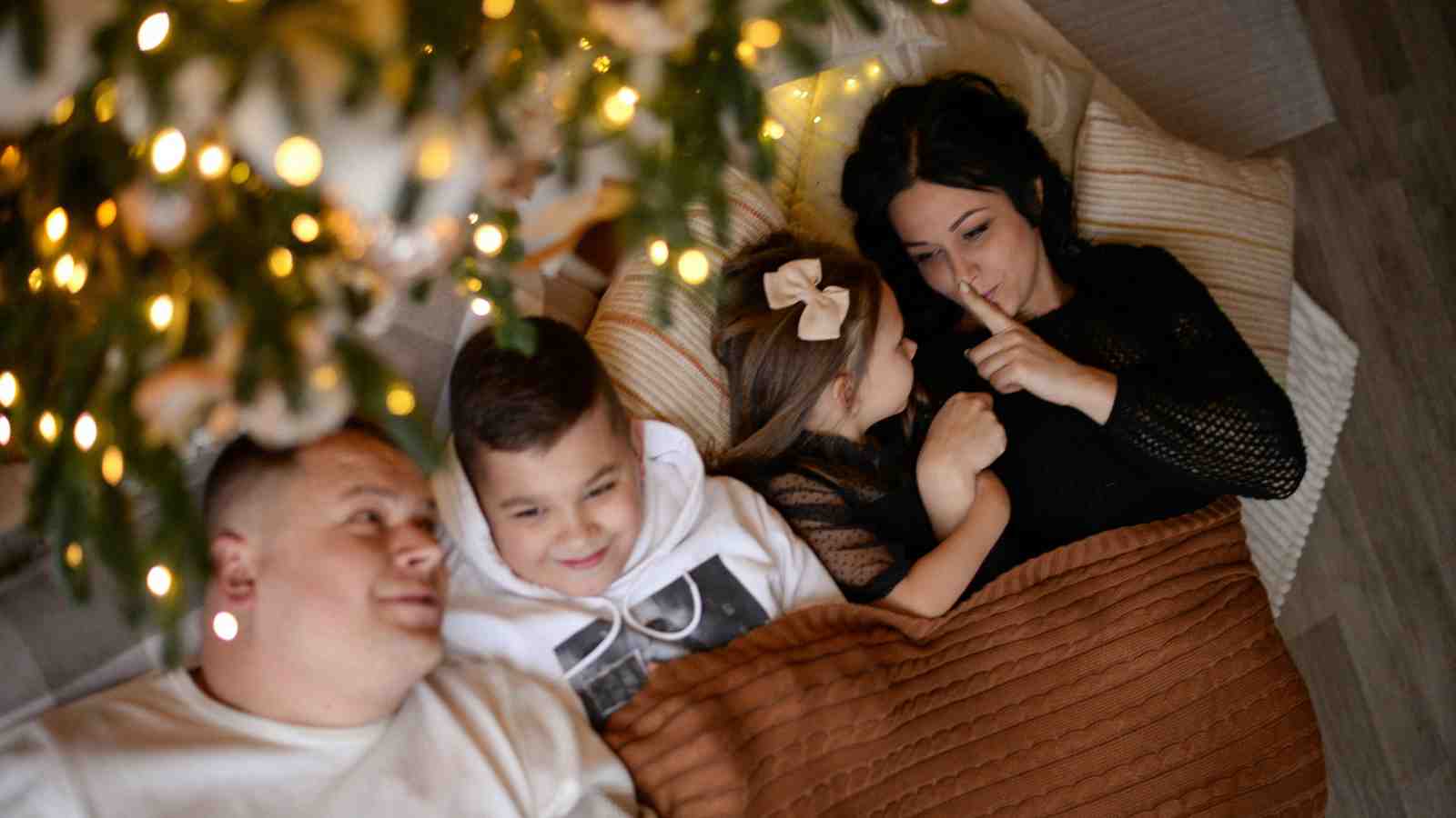 A family of four lies together under a blanket near a lit Christmas tree, with the mother holding a finger to her lips, signaling for quiet.